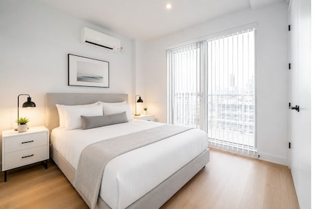 A modern bedroom with a bed, bedside tables, a window with blinds, and an air conditioning unit.
