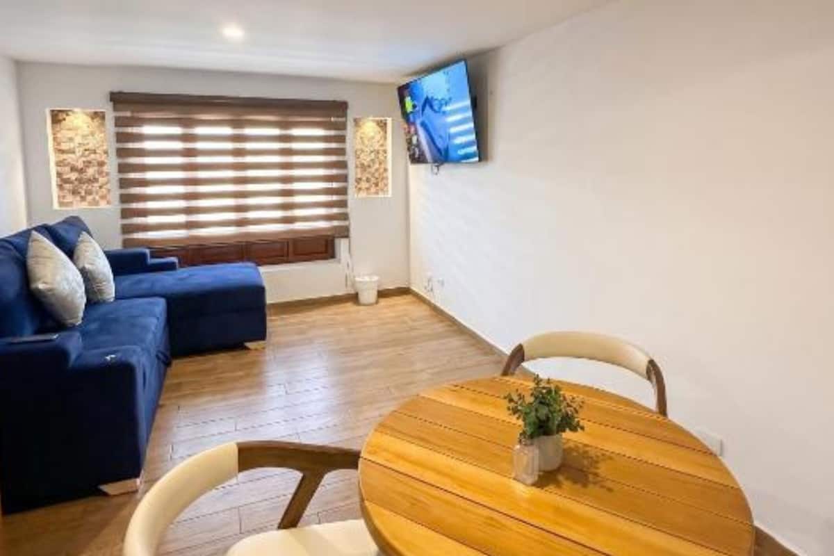 A living room with a blue sofa, a wooden dining table with chairs, a TV mounted on the wall, and a window with blinds.