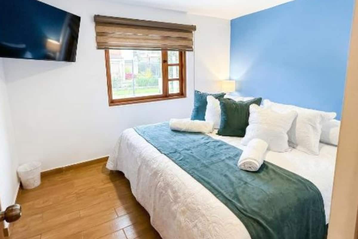 A bedroom with a bed, a TV mounted on the wall, a window with blinds, and a wooden floor.