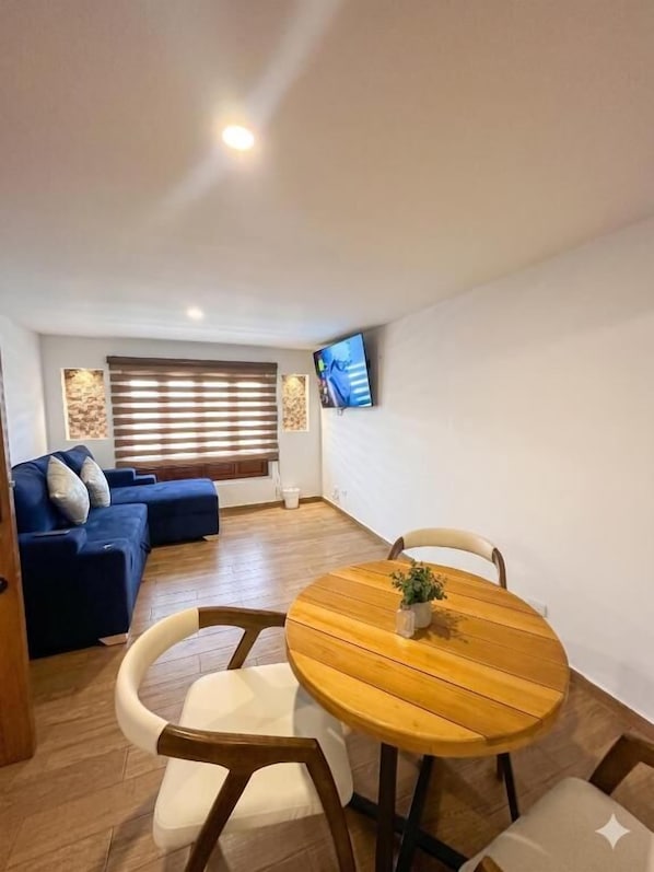 A living room with a round wooden table, two white cushioned chairs, a blue sofa, a flat-screen TV mounted on the wall, and a window with blinds.