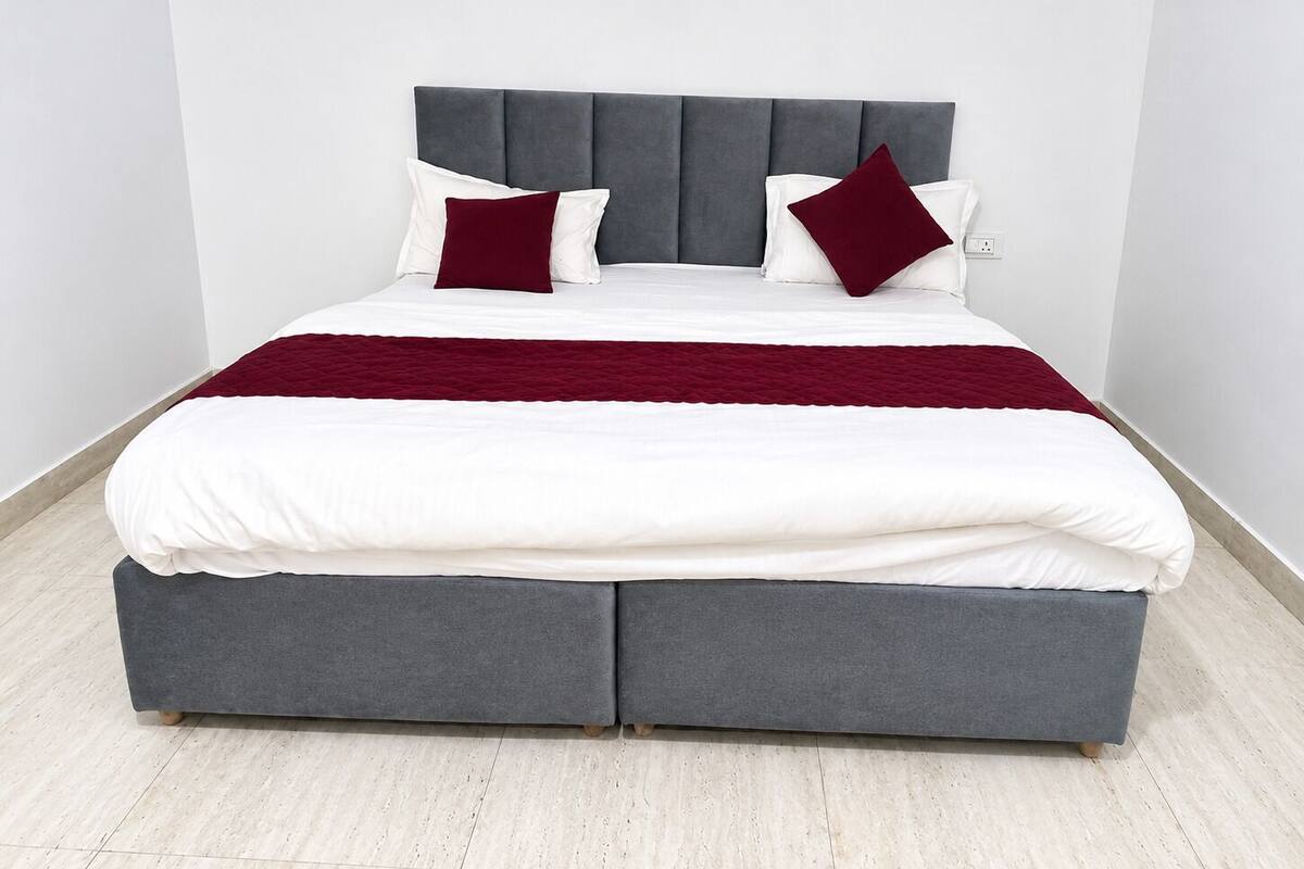 Una cama con cabecera tapizada en gris, ropa de cama blanca y dos almohadas rojas.