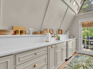 A kitchen with a white countertop, wooden utensils, a clock, and a window with a view of greenery.