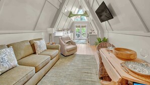 A cozy loft bedroom with a high ceiling, a large window, a wooden dining table, and a comfortable sofa.