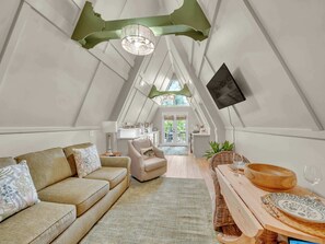 A cozy loft bedroom with a high ceiling, a large window, a wooden dining table, and a comfortable sofa.