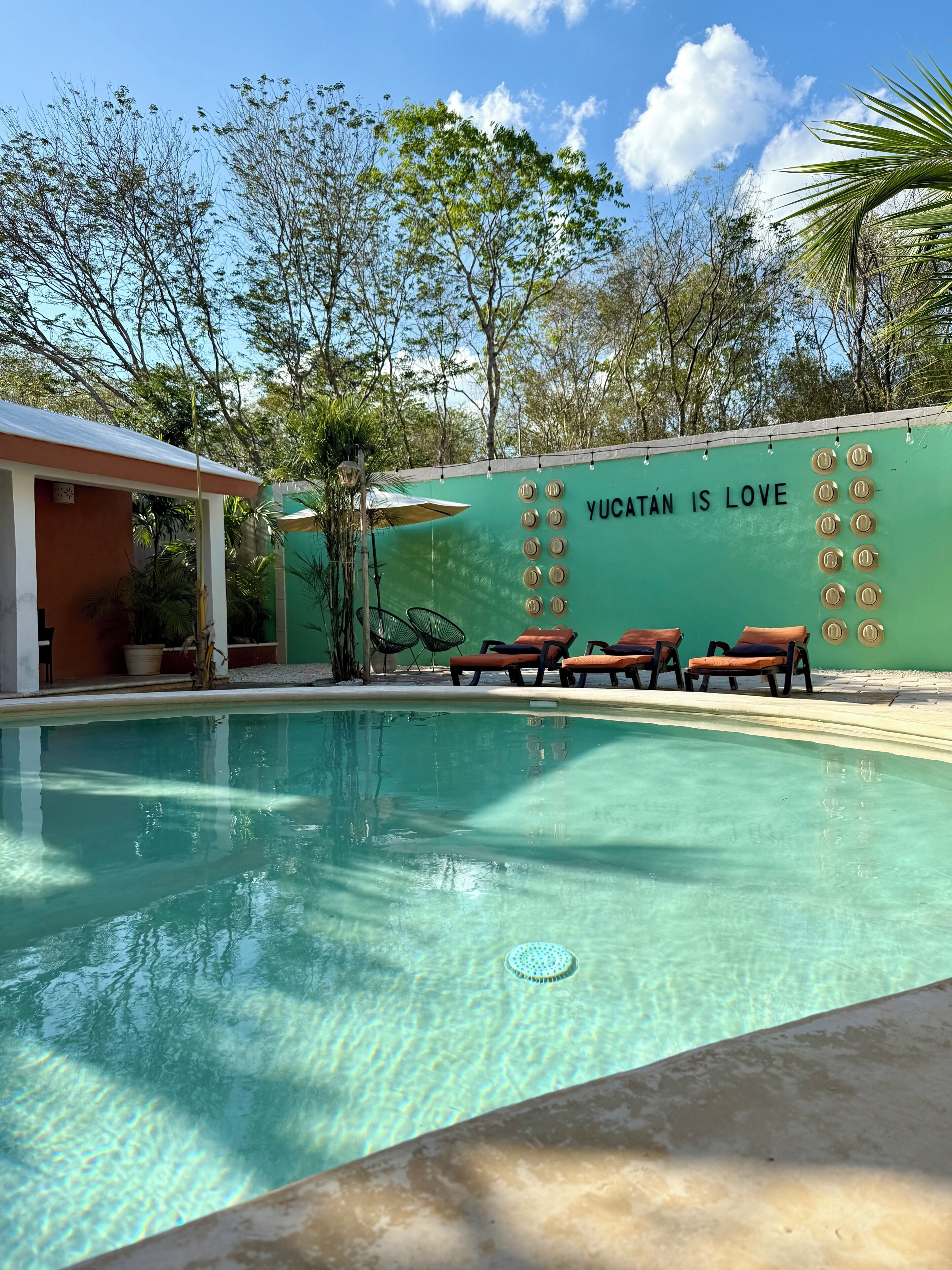 Una piscina con sillas de descanso, una pared con el texto 'YUCATÁN IS LOVE' y árboles circundantes.