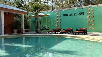 Una piscina con sillas de descanso, una pared con el texto 'YUCATÁN IS LOVE' y árboles circundantes.