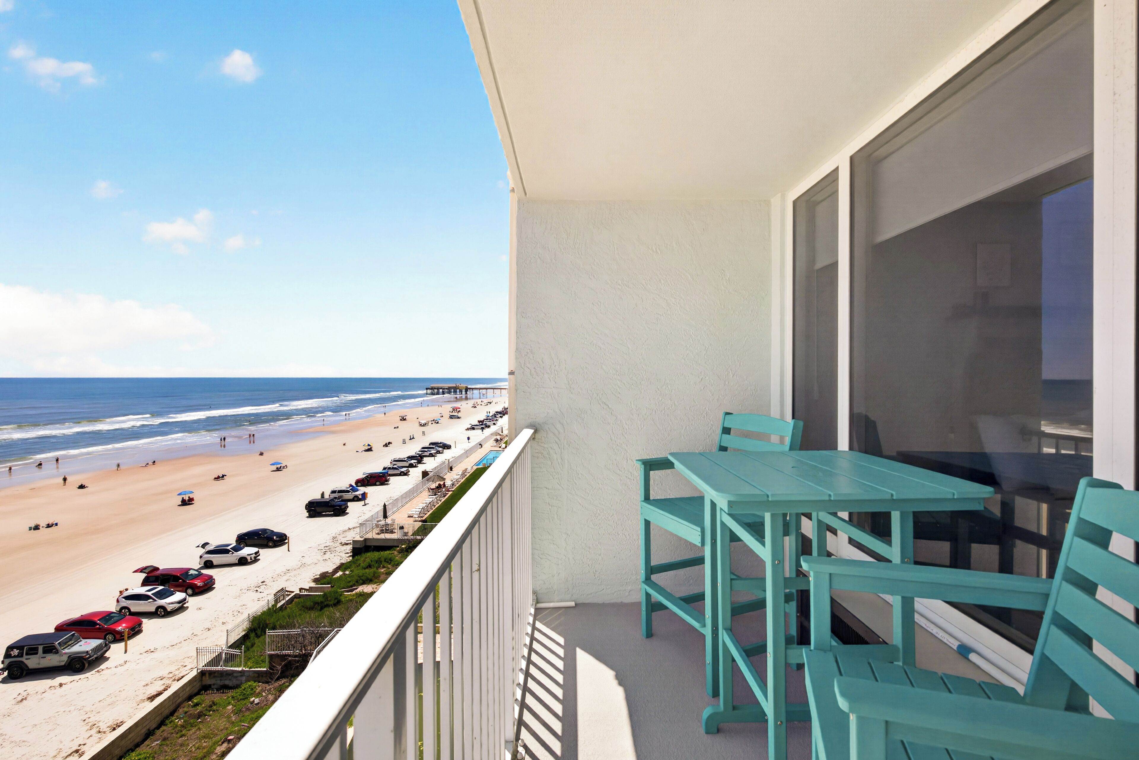Een balkon met turquoise stoelen en een tafel met uitzicht op een strand waar auto's geparkeerd staan en mensen zijn.