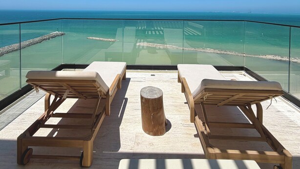 A balcony with two wooden lounge chairs and a small wooden stool, overlooking a clear blue sea and a rocky shoreline. - Waterfront penthouse in Yucalpetén Resort Marina (Progreso)