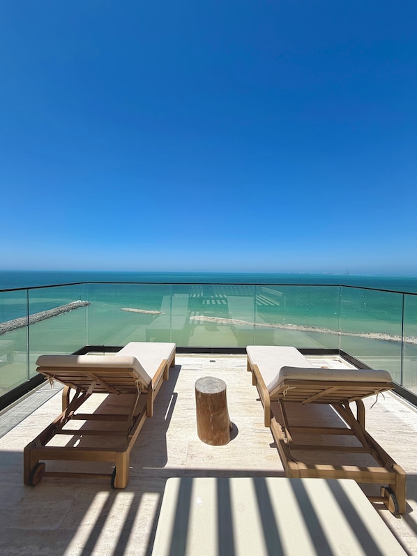 Ein Balkon mit zwei Holzliegen und einem kleinen Hocker, mit Blick auf ein klares blaues Meer und eine felsige Küste.