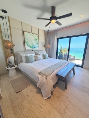 Ein Schlafzimmer mit einem großen Bett, einem Deckenventilator, einem Nachttisch, einer Stehlampe, einem gerahmten Bild und Blick auf das Meer durch ein großes Fenster.