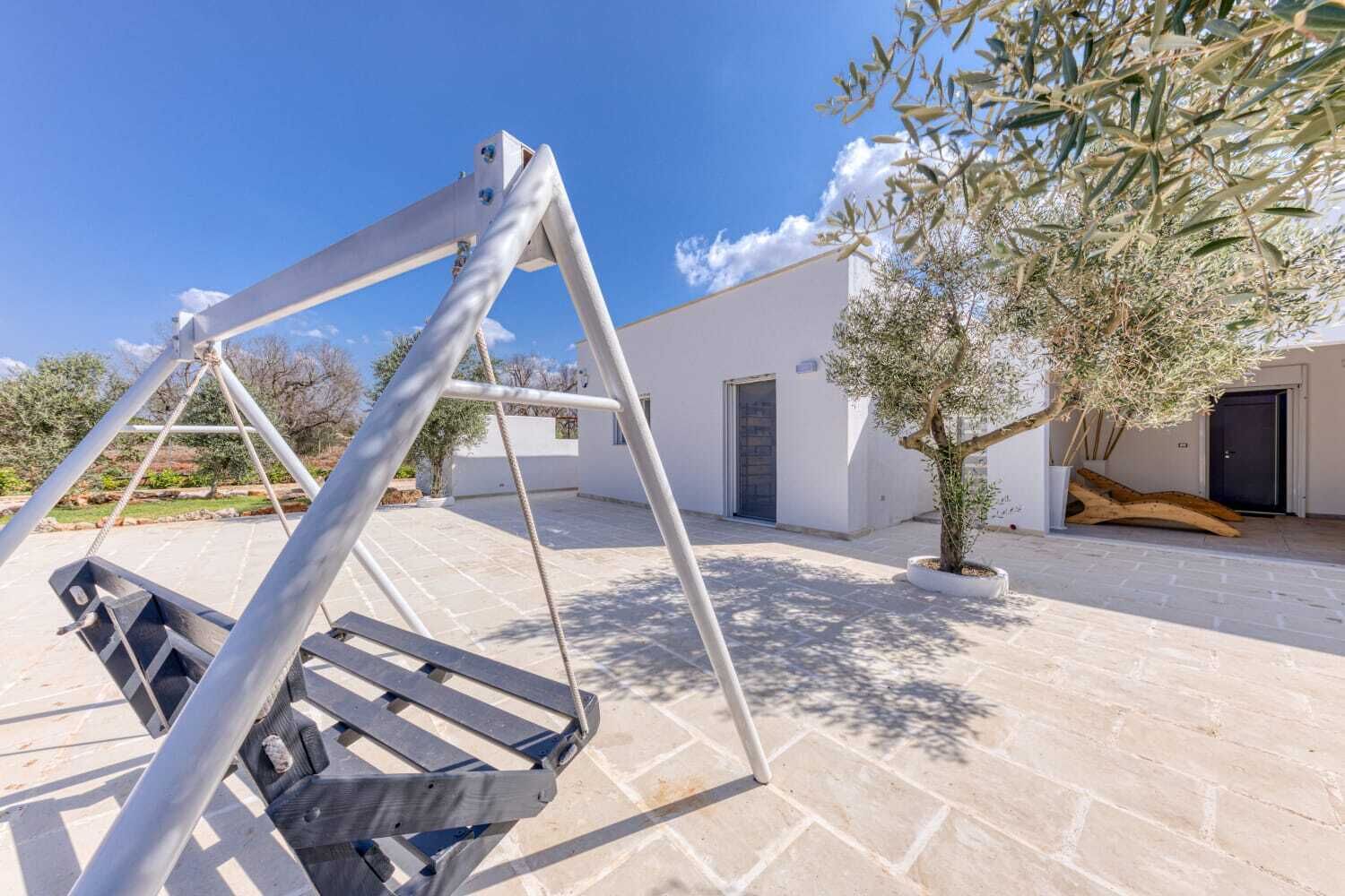 A patio with a swing set, a potted olive tree, and a white building with a door.