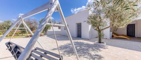 A patio with a swing set, a potted olive tree, and a white building with a door.