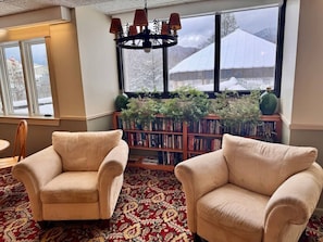 Ein Zimmer mit zwei beigen Sesseln, einem gemusterten Teppich, einem Bücherregal und Blick auf schneebedeckte Gebäude durch große Fenster.