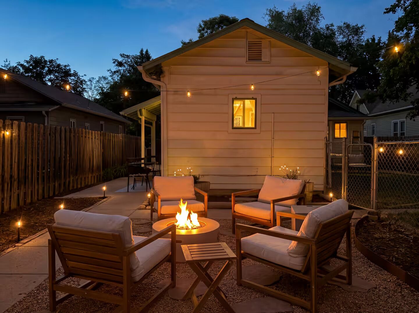 A cozy outdoor seating area with a fire pit, surrounded by wooden furniture and string lights.
