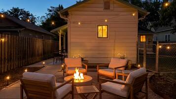 A cozy outdoor seating area with a fire pit, surrounded by wooden furniture and string lights.
