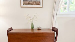 Une salle à manger avec une table en bois, des chaises, un vase de fleurs, un tableau encadré au mur et une fenêtre avec des rideaux.