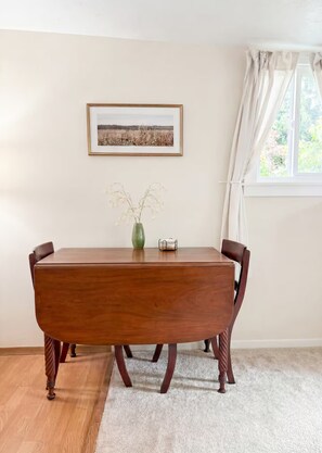 A dining room with a wooden table, chairs, a vase with flowers, a framed picture on the wall, and a window with curtains.