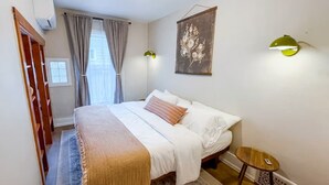 Une chambre à coucher avec un lit, une petite table ronde, une lampe fixée au mur et une fenêtre avec des rideaux.
