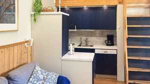 Eine kompakte Küche mit Spüle, Herd und Kaffeemaschine, eine kleine Sitzecke mit Sofa und Kissen sowie ein Schlafoberschlaf über der Küche.