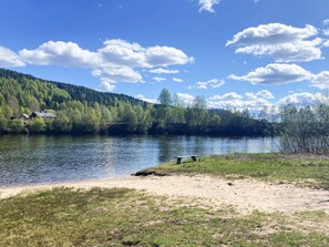 A serene lakeside view with a wooden bench, a sandy shore, and a forested hill in the background.