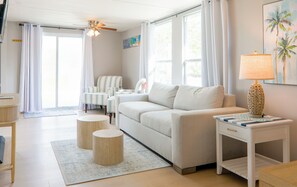 Ein Wohnzimmer mit einer Couch, zwei Beistelltischen, einer Lampe, einem Deckenventilator und einem fenster mit Vorhängen.