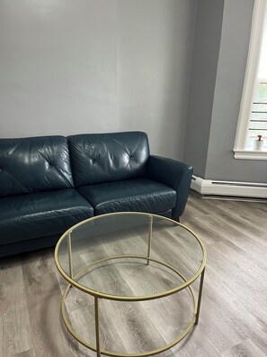 Ein dunkelblaues Ledersofa, ein runder Glastisch mit goldenen Beinen und eine graue Wand mit einem Fenster.