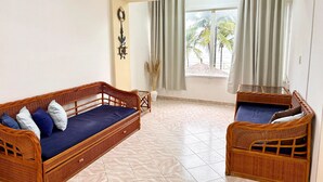 Una habitación con dos camas de mimbre, un ventanal con cortinas, un adorno en la pared y piso de baldosas.