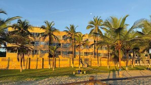 Un edificio de varios pisos con fachada de ladrillo, rodeado de palmeras y una zona arenosa con una cerca de madera.