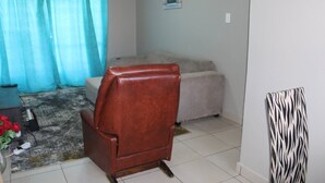 A living room with a brown leather chair, a beige sofa, a patterned wall hanging, and a window with a view. - Beautiful City Centred Penthouse (Bloemfontein)