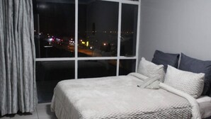 A bedroom with a large window, a bed with white bedding, and a view of a cityscape at night. - Beautiful City Centred Penthouse (Bloemfontein)