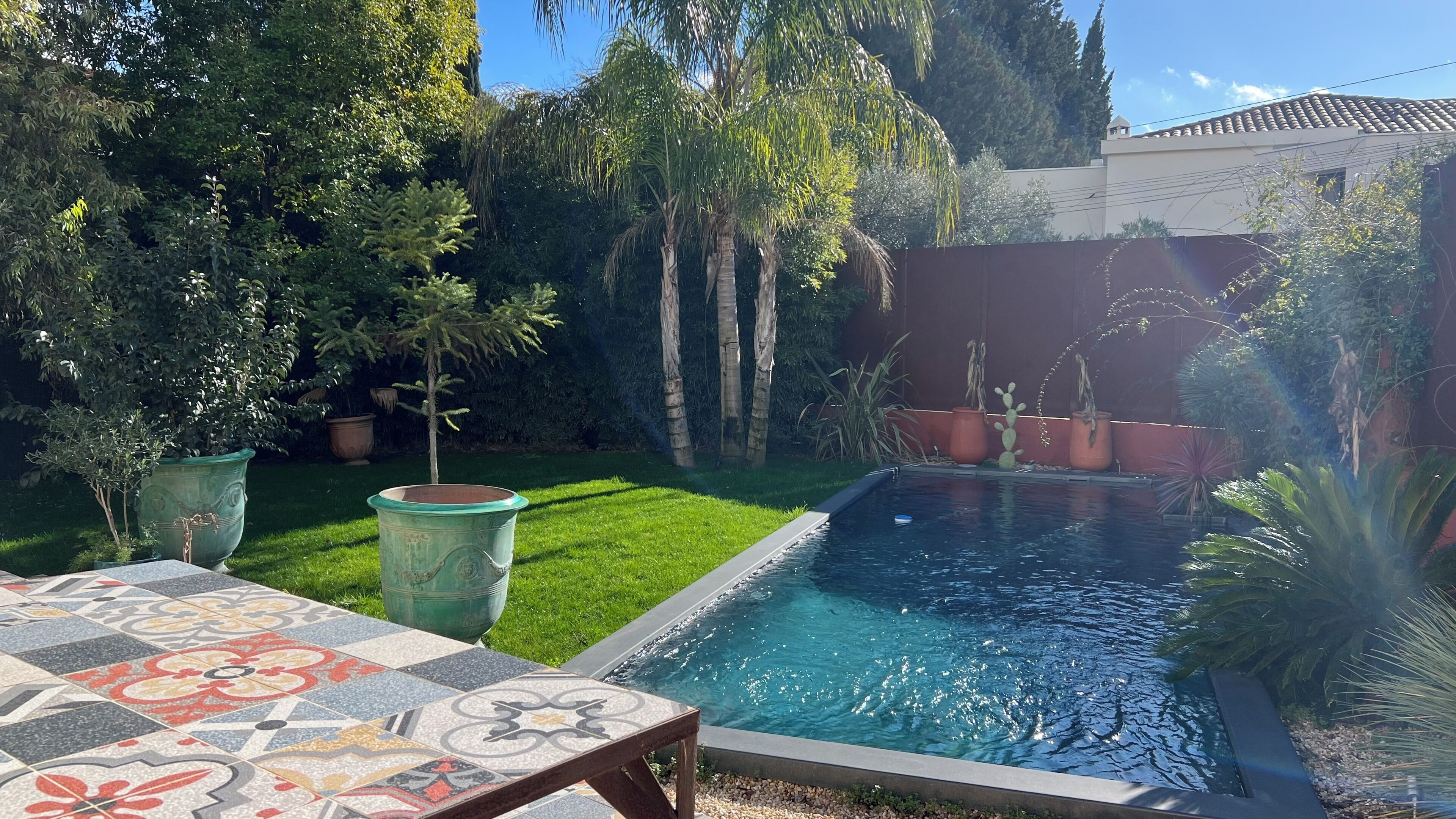 A swimming pool with a tiled deck, surrounded by lush greenery and potted plants.