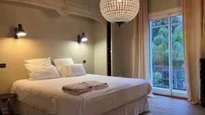 A bedroom with a large bed, two wall-mounted lights, a chandelier, and a view of greenery through a window. - 12345677890 (Montpellier)
