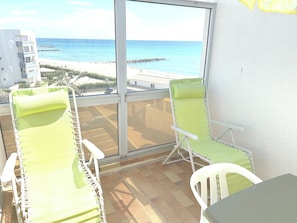 A balcony with two green lounge chairs, a white table, and a view of the sea and beach. - BRIGHT STUDIO WITH TERRACE FACING THE SEA AND MARINA (Le Barcarès)