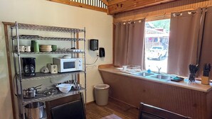 A kitchen with a wooden table, chairs, a metal shelf with kitchenware, a microwave, and a window with curtains.