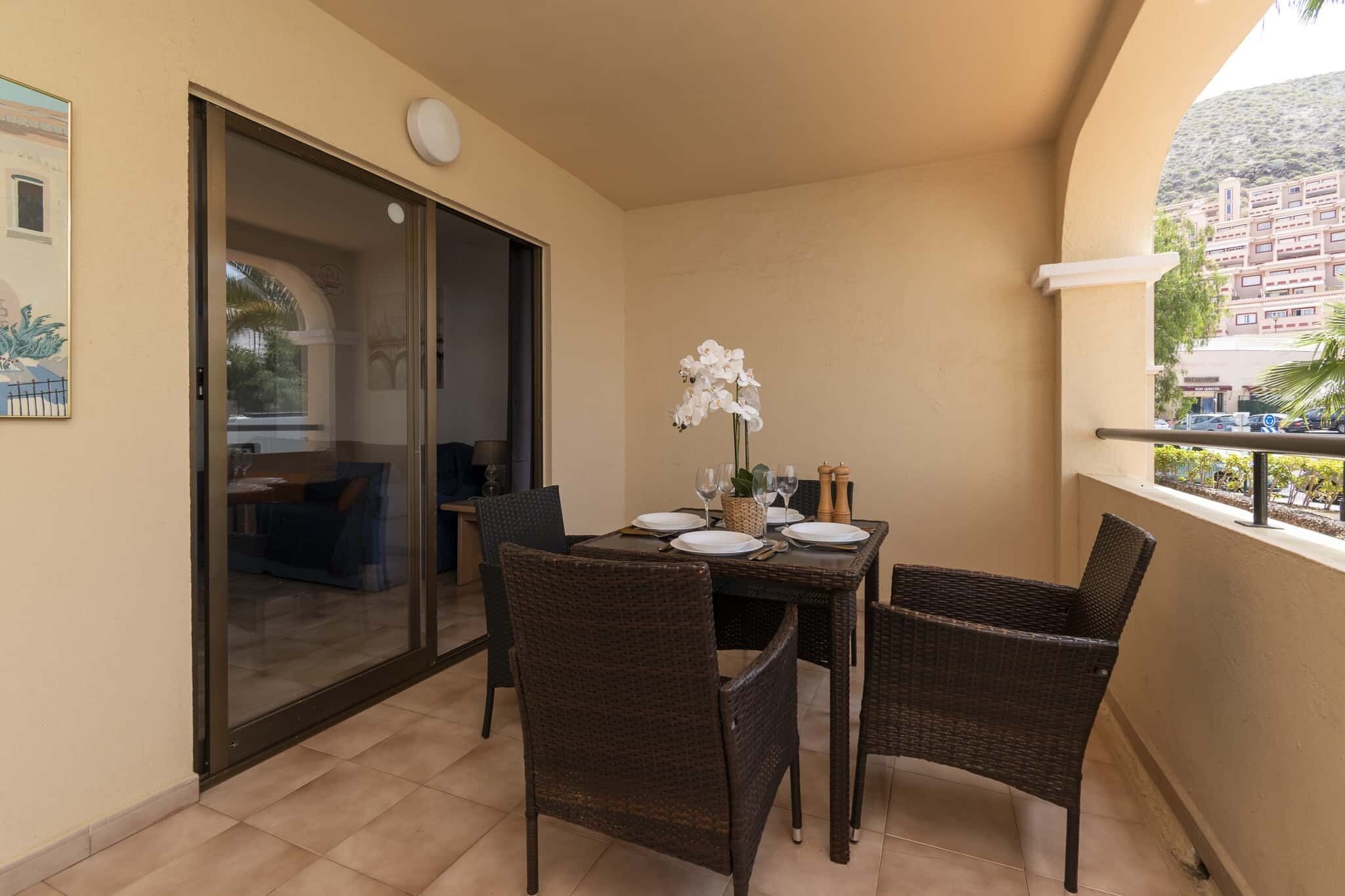 A balcony with a dining table set for four, wicker chairs, a potted orchid, and a sliding glass door leading to an outdoor area.