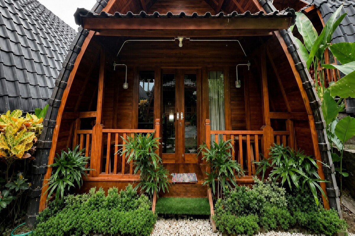 Ein Holzhaus mit einem steilen Dach, einer Veranda mit Geländern und einem kleinen Vorgarten mit verschiedenen Pflanzen.