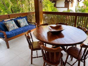 Eine Holzterrasse mit einem runden Tisch und Stühlen, ein blaues Sofa mit gemusterten Kissen und Blick auf Grünflächen.