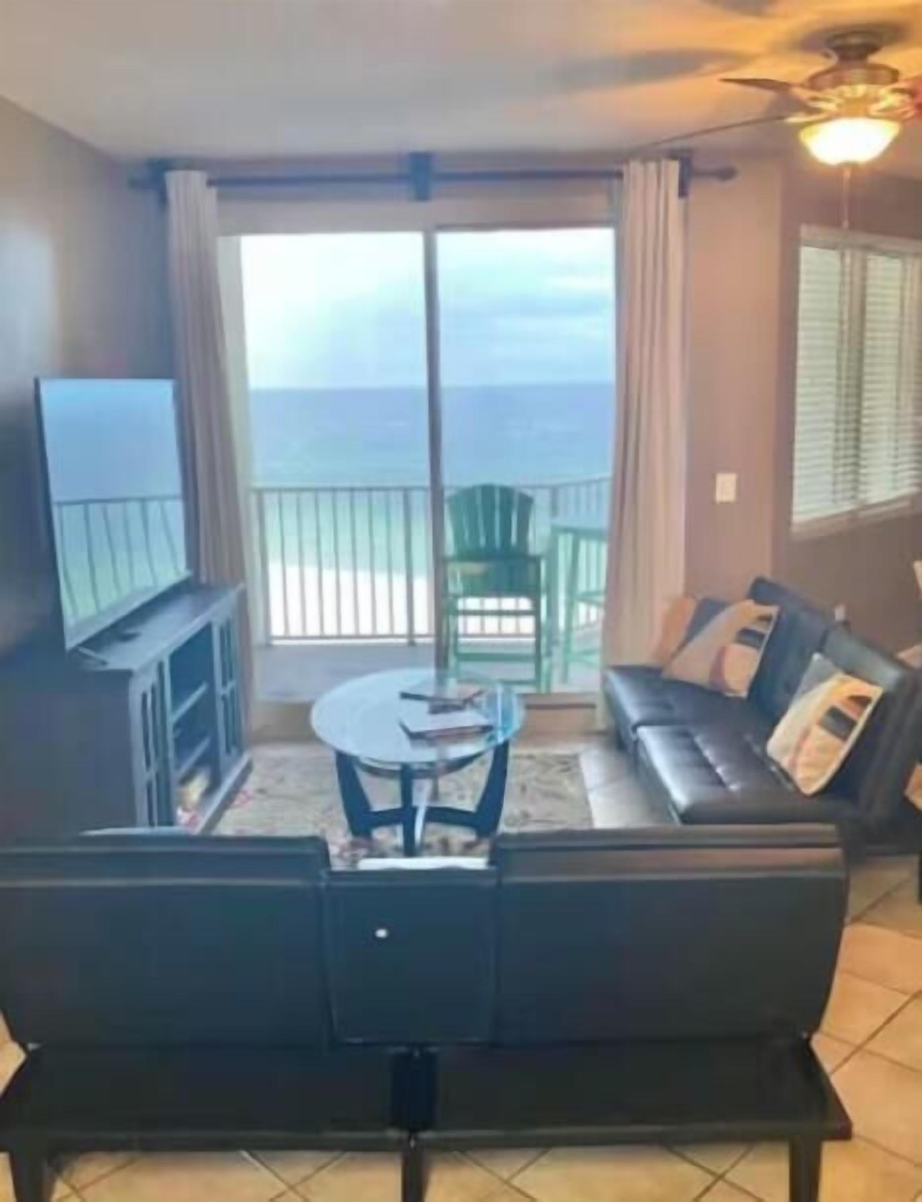 Een woonkamer met een bank, een televisie, een tafel en een balkon met uitzicht op de oceaan.