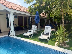 A swimming pool with two white lounge chairs and a blue umbrella, surrounded by greenery and a tiled-roof building. - Family house with swimming pool 5 minutes from shopping centers, San Miguel (San José Quelepa)