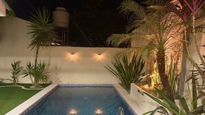 Une piscine rectangulaire dont le bord est recouvert de carreaux, entourée de plantes et bordée d’un mur blanc.
