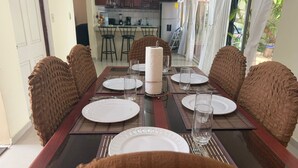 Une table à manger dressée pour six personnes, avec des chaises en osier, une bougie au centre et une cuisine en arrière-plan.