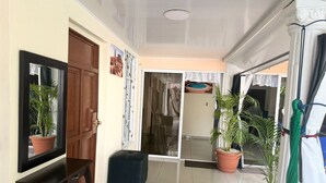 Un couloir avec une porte en bois, un miroir, une petite table, une valise, des plantes en pot et une porte-fenêtre coulissante donnant sur une pièce.