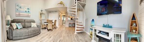 Ein Wohnzimmer mit einer gebogenen grauen Couch, Korbsesseln, einer weißen Wendeltreppe, einem an der Wand montierten Fernseher und einem kleinen Holzregal mit Dekoration.