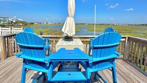 Una terraza de madera con dos sillas Adirondack azules y una mesa cubierta, con vistas a un paisaje costero.