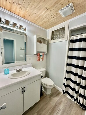 A bathroom with a wooden ceiling, a white sink with a soap dispenser, a toilet, and a striped shower curtain. - Seas the Day Beach Retreat - Waterfront Views + Hot Tub + Kayaks (North Topsail Beach)