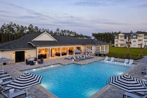 A swimming pool with lounge chairs and umbrellas, a covered patio area, and a building with a sloped roof. - Near Amelia Island. KingBed. Pool. Pickle Ball Gym (Yulee)