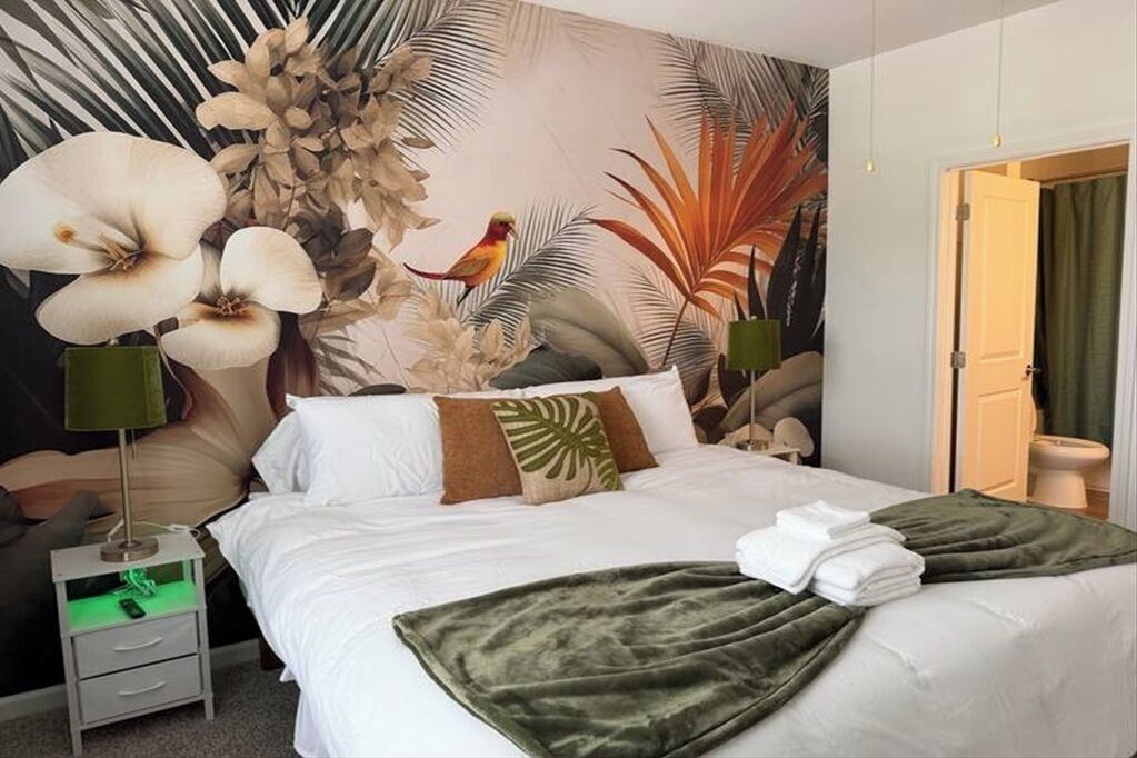 Ein Hotelzimmer mit Bett, Nachttisch, grüner Decke und einem Wandbild im tropischen Stil.