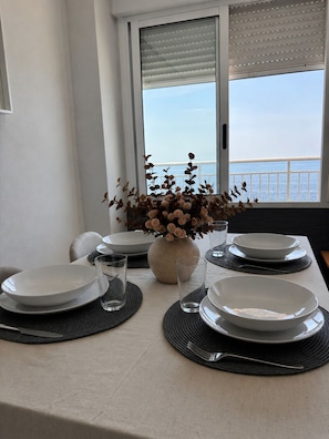 Une table Ă manger dressĂ©e avec des assiettes, des verres et un vase de fleurs, offrant une vue sur la mer par la fenĂȘtre.