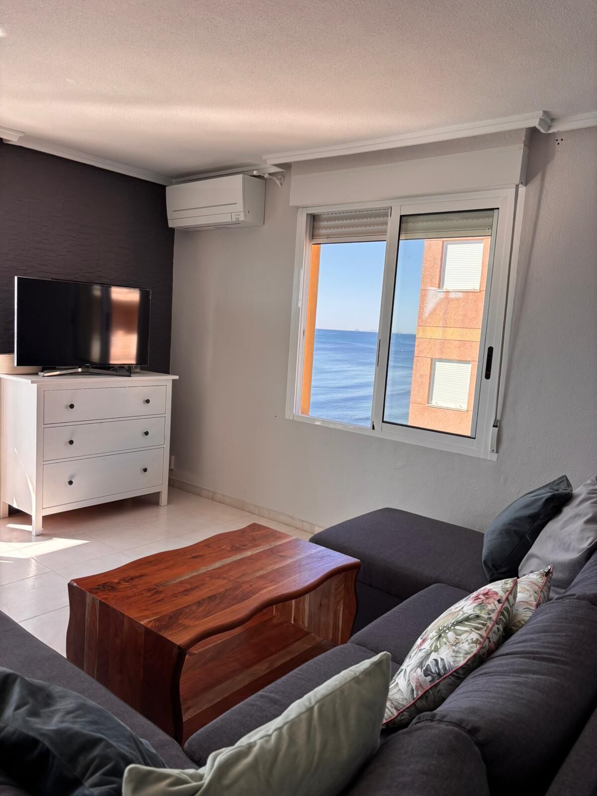 Una sala de estar con un sofá, una mesa de centro de madera, un televisor sobre un mueble blanco y una ventana con vista al mar.