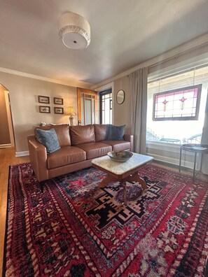 Ein Wohnzimmer mit einem braunen Ledersofa, einem gemusterten Teppich, einem kleinen Holztisch und einem großen Fenster mit Blick.